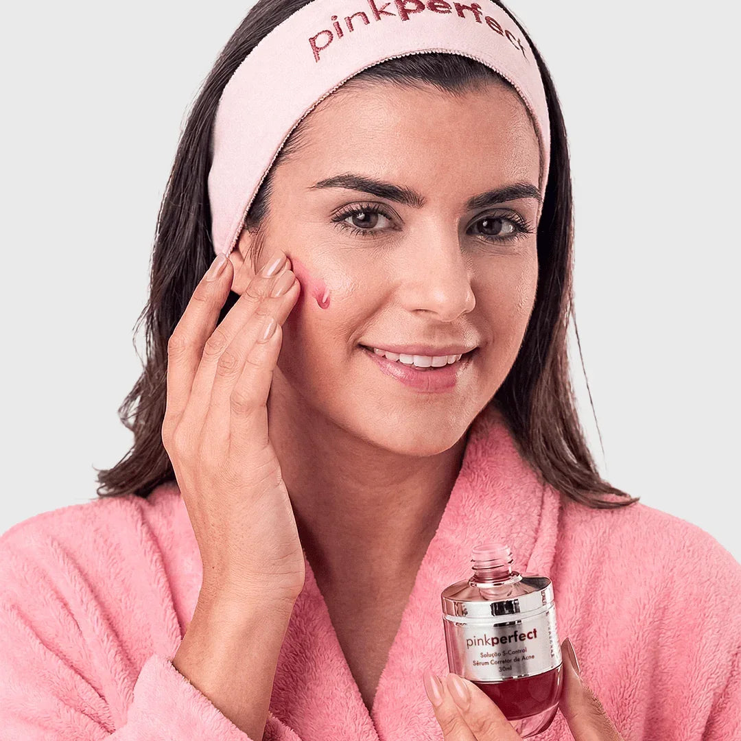 Mulher aplicando sérum facial para acne adulta, usando faixa de cabelo e roupão rosa